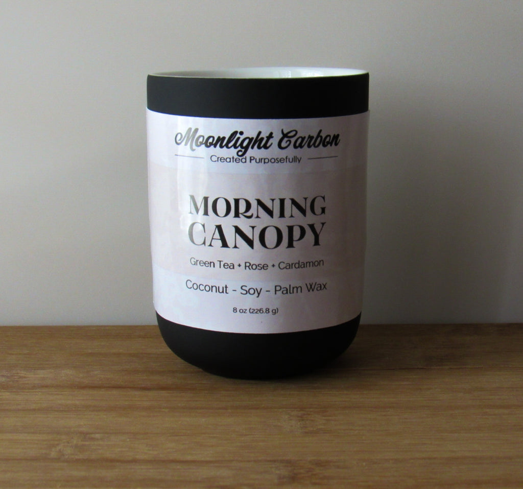 Coconut, soy, and palm wax candle labeled 'Morning Canopy' by Moonlight Carbon on a wooden surface with a neutral background. Fragrance notes include green tea, rose, and cardamon. 