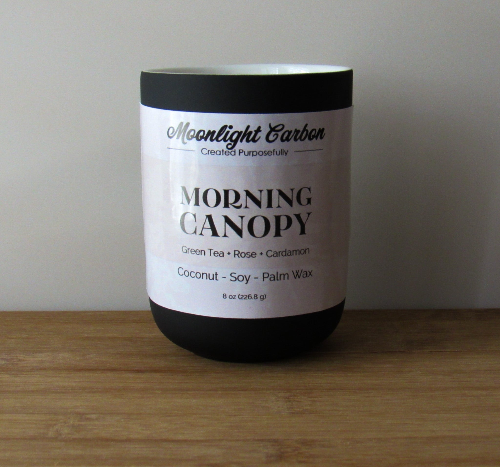 Coconut, soy, and palm wax candle labeled 'Morning Canopy' by Moonlight Carbon on a wooden surface with a neutral background. Fragrance notes include green tea, rose, and cardamon. 