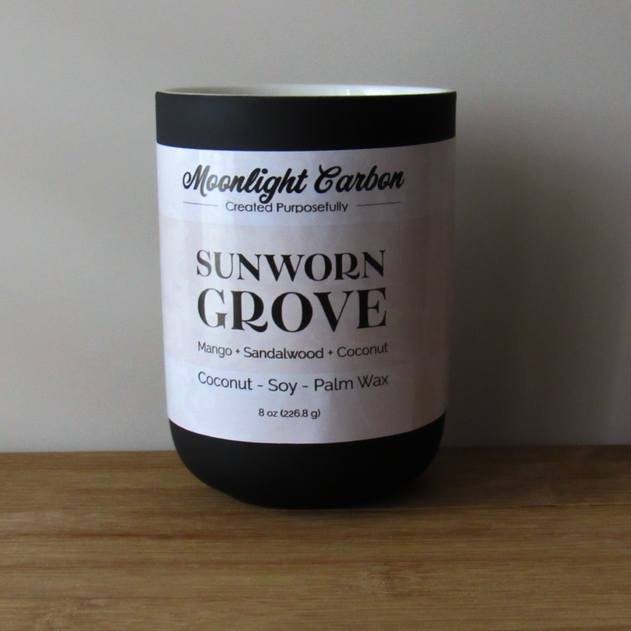 Coconut, soy, and palm wax candle labeled 'Sunworn Grove' by Moonlight Carbon on a wooden surface with a neutral background. Fragrance notes include mango, sandalwood, and coconut. 
