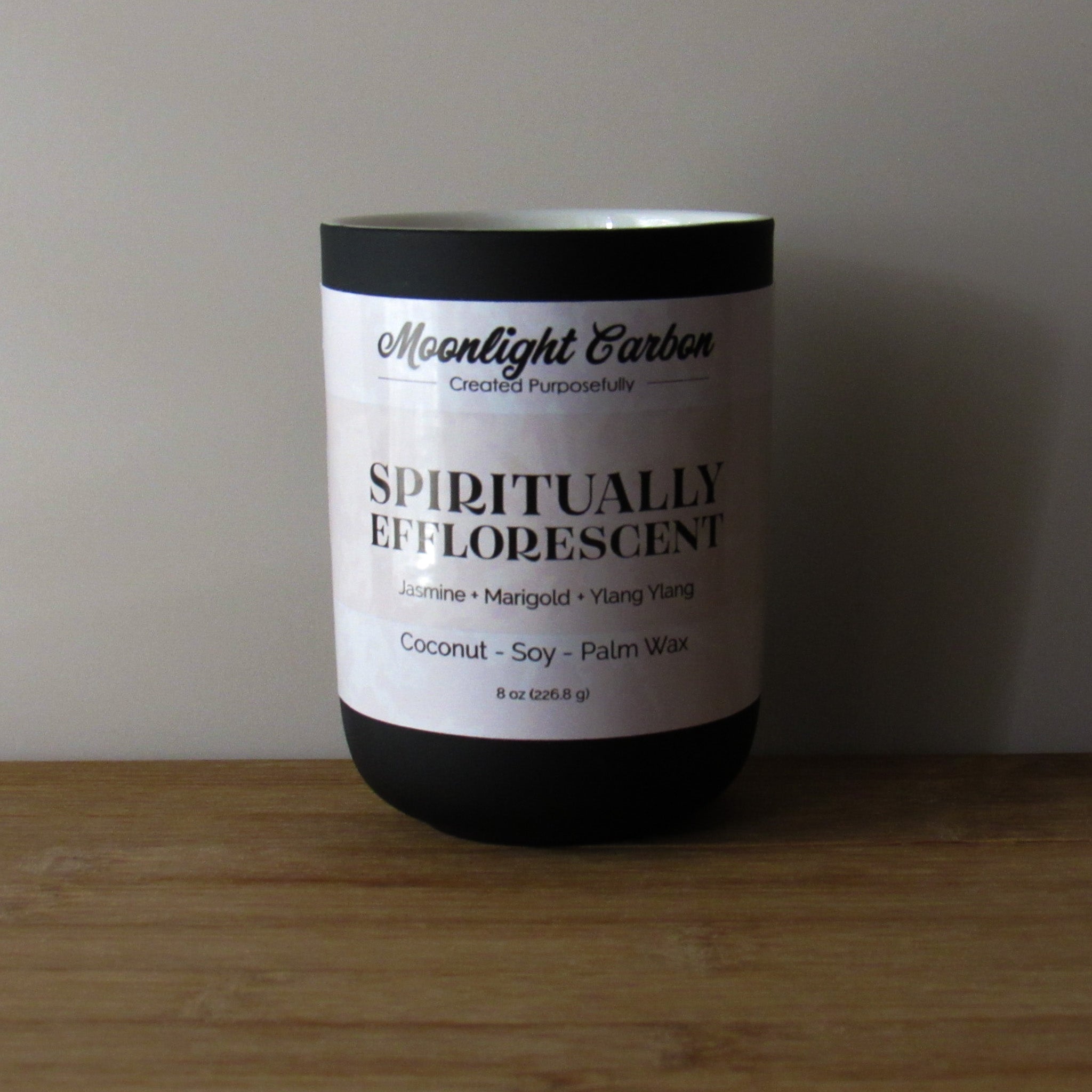 Coconut, soy, and palm wax candle labeled 'Spiritually Efflorescent' by Moonlight Carbon on a wooden surface with a neutral background. Fragrance notes include jasmine, marigold, and ylang ylang. 