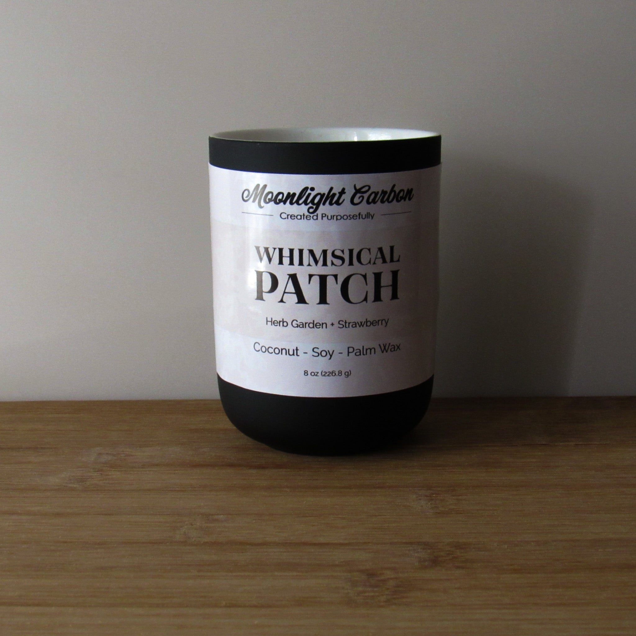 Candle labeled 'Whimsical Patch' on a wooden surface and gray background. Fragrance notes include herb garden and strawberries.