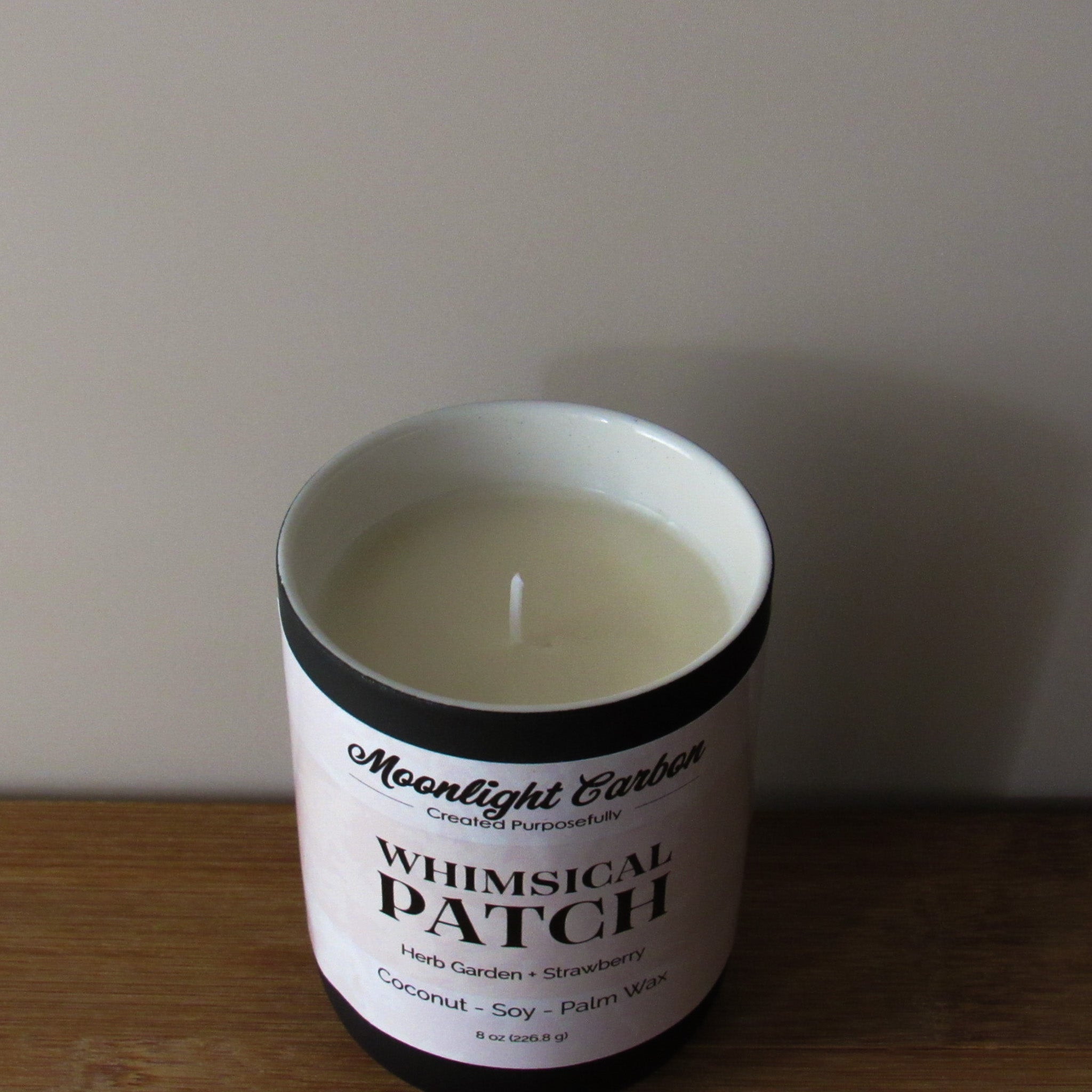 Candle in a container labeled 'Whimsical Patch' on a wooden surface with a neutral background. Camera zoomed into top view of the candle showing the coconut, soy, palm wax blend and cotton wick.