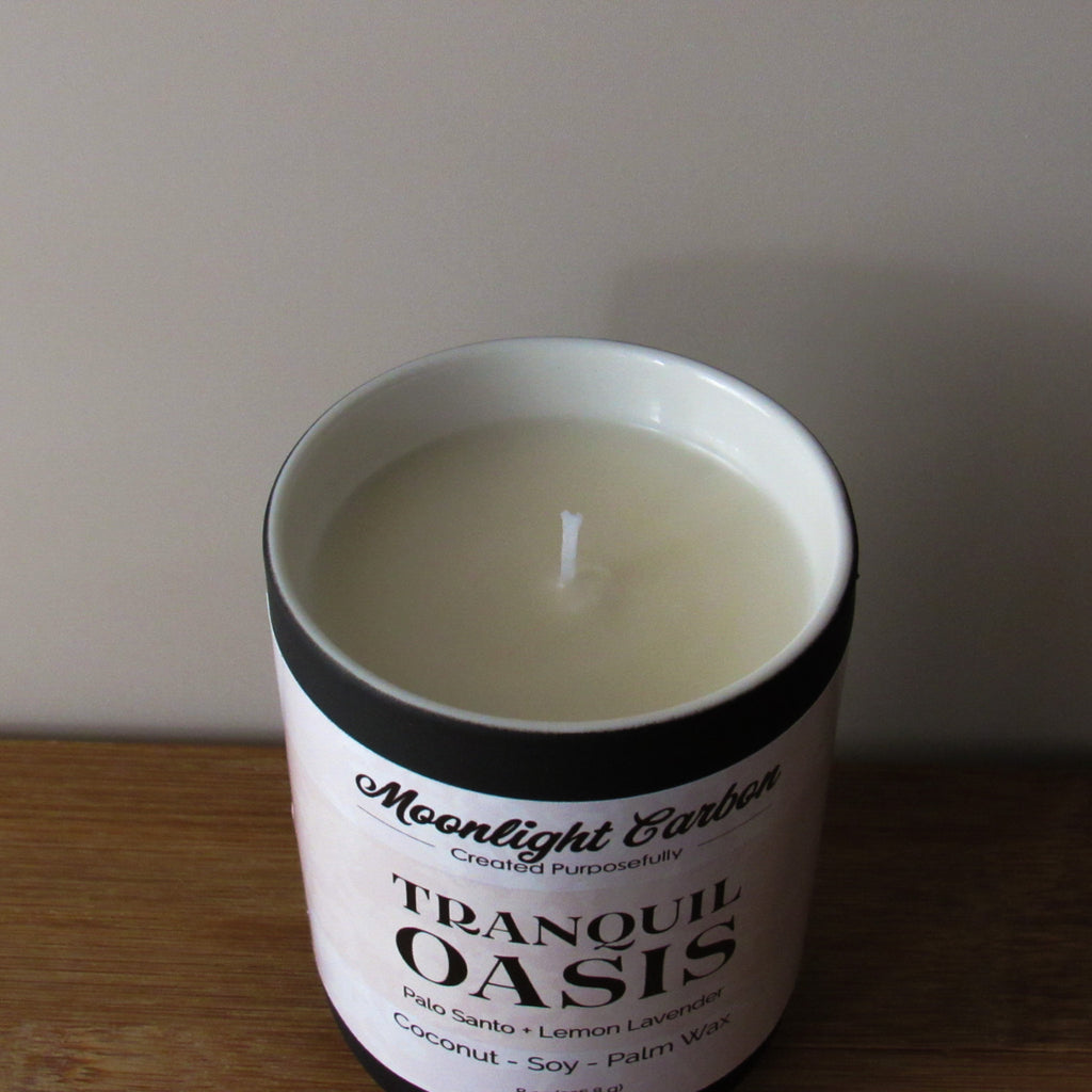 Candle in a container labeled 'Tranquil Oasis' by Moonlight Carbon on a wooden surface. zoomed into top view of candle showing coconut, soy, palm wax blend and cotton wick.