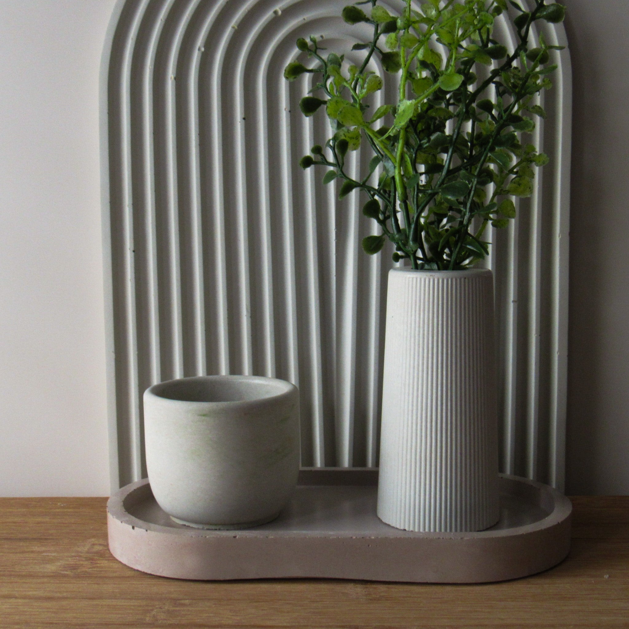 Two concrete home goods. A mini candle and a mini vase on a concrete tray with a concrete gray rainbow ridged tray in background.