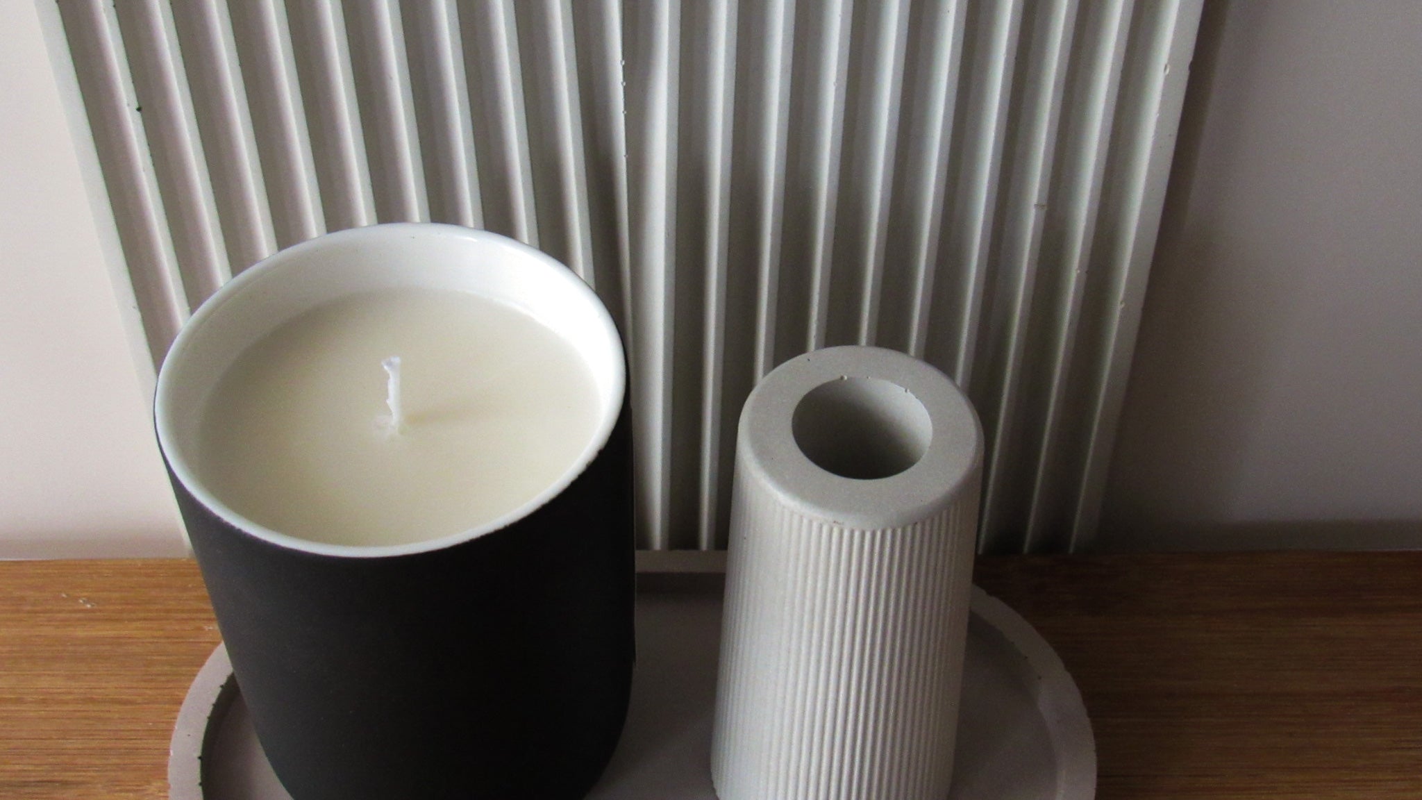Candle in a black ceramic jar and a gray concrete mini vase on a wooden surface with a large rainbow shape ridged tray behind.