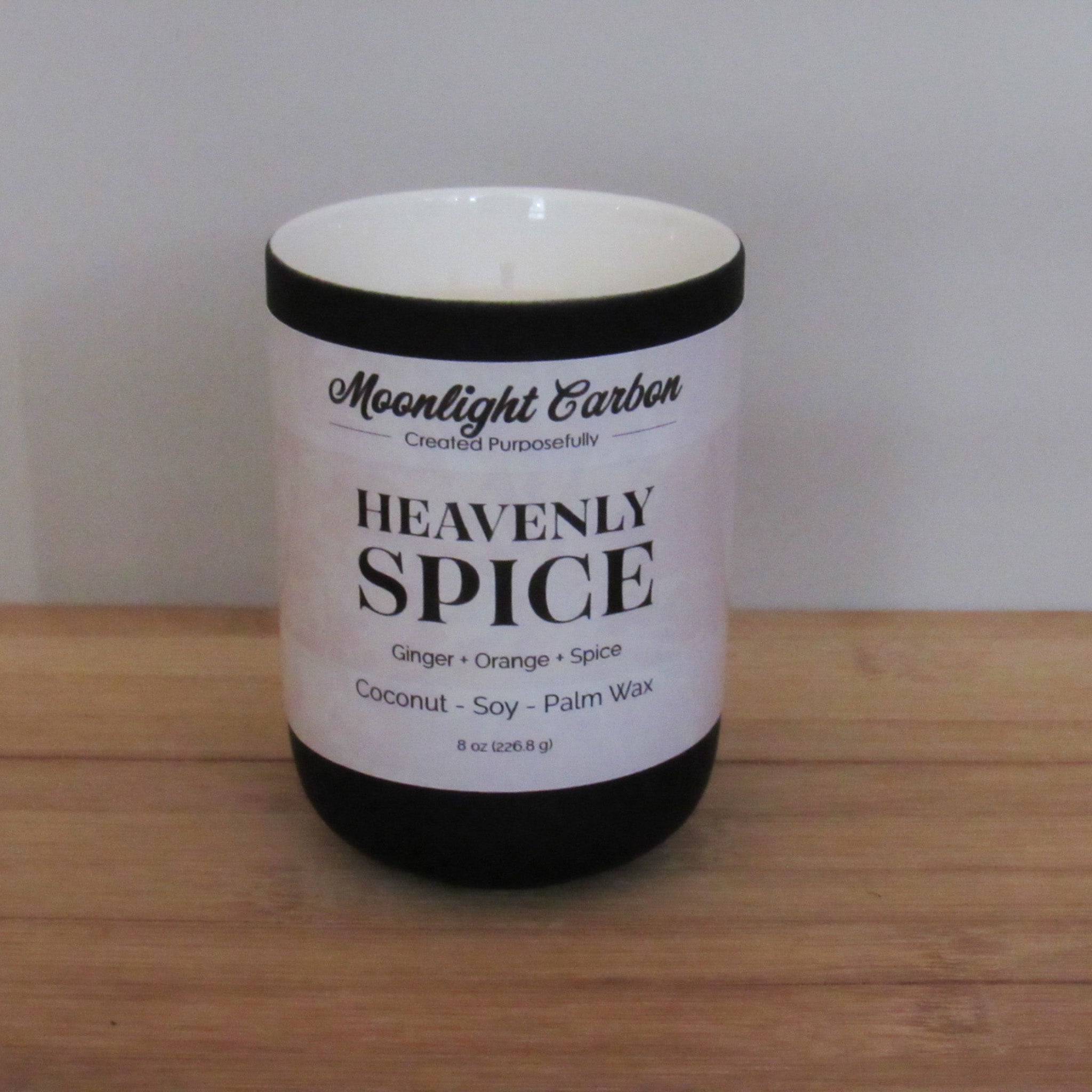 Candle labeled 'Heavenly Spice' by Moonlight Candles on a wooden surface with a white background, In a black ceramic candle jar. Fragrance notes include ginger, orange, and spice.