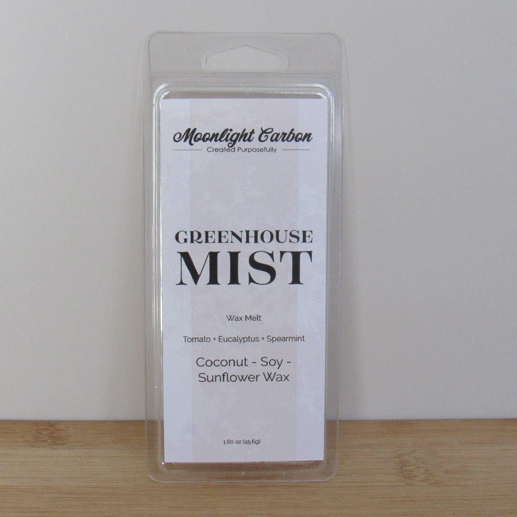 Packaged wax melt labeled 'Greenhouse Mist' by Moonlight Carbon on a wooden surface with a white background. Fragrance notes include tomato, eucalyptus, and spearmint.