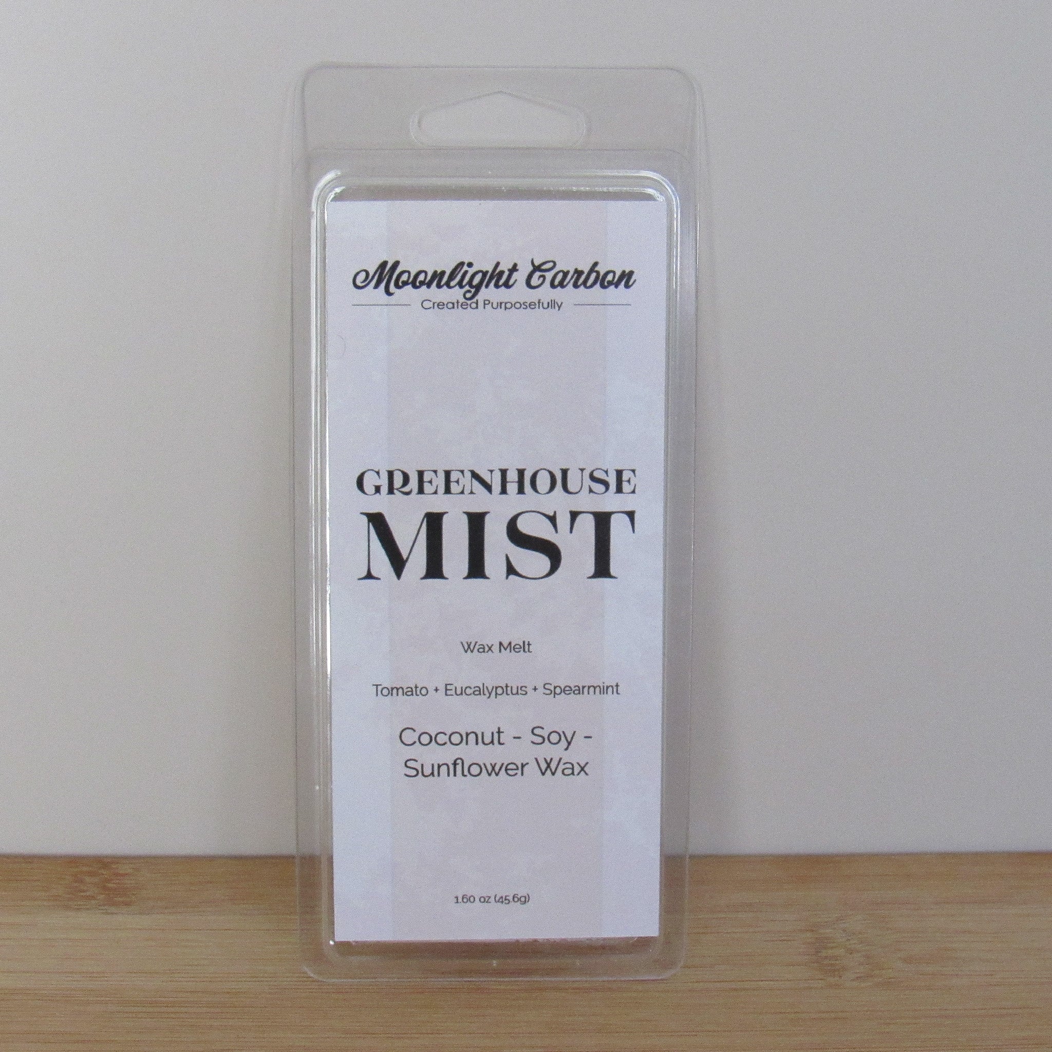 Packaged wax melt labeled 'Greenhouse Mist' by Moonlight Carbon on a wooden surface with a white background. Fragrance notes include tomato, eucalyptus, and spearmint.