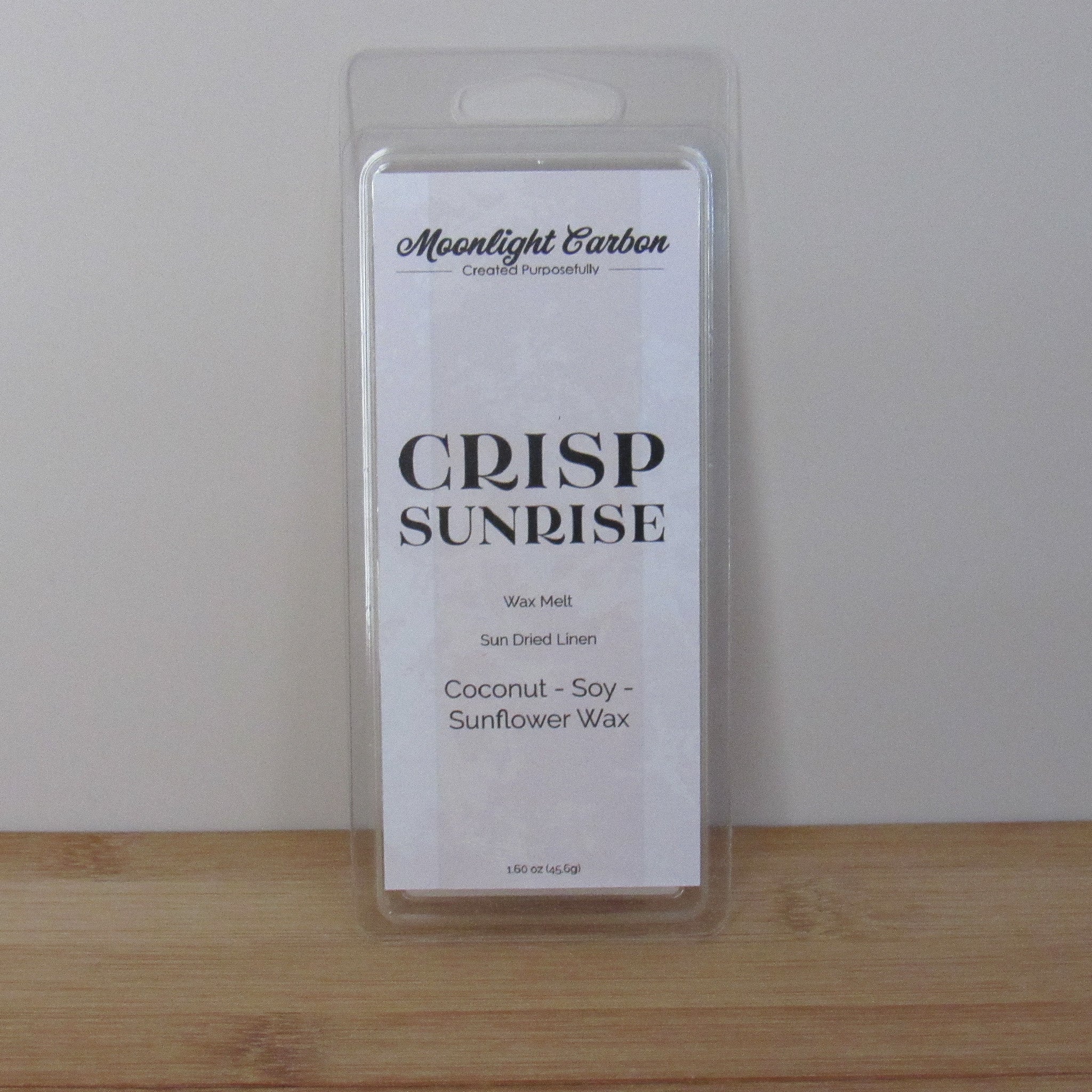 Packaged wax melt labeled 'Crisp Sunrise' by Moonlight Carbon on a wooden surface with a white background. Fragrance notes include fresh laundry.