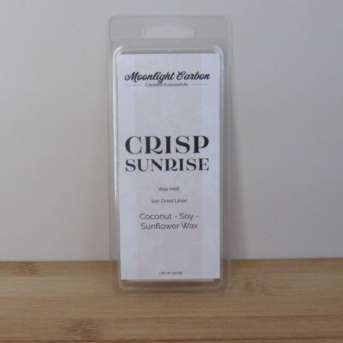 Packaged wax melt labeled 'Crisp Sunrise' by Moonlight Carbon on a wooden surface with a white background. Fragrance notes include fresh laundry.