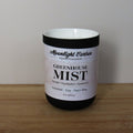 Candle with a labeled 'Greenhouse Mist' on a wooden surface and plain background. Fragrance notes include tomato, eucalptus, and spearmint.
