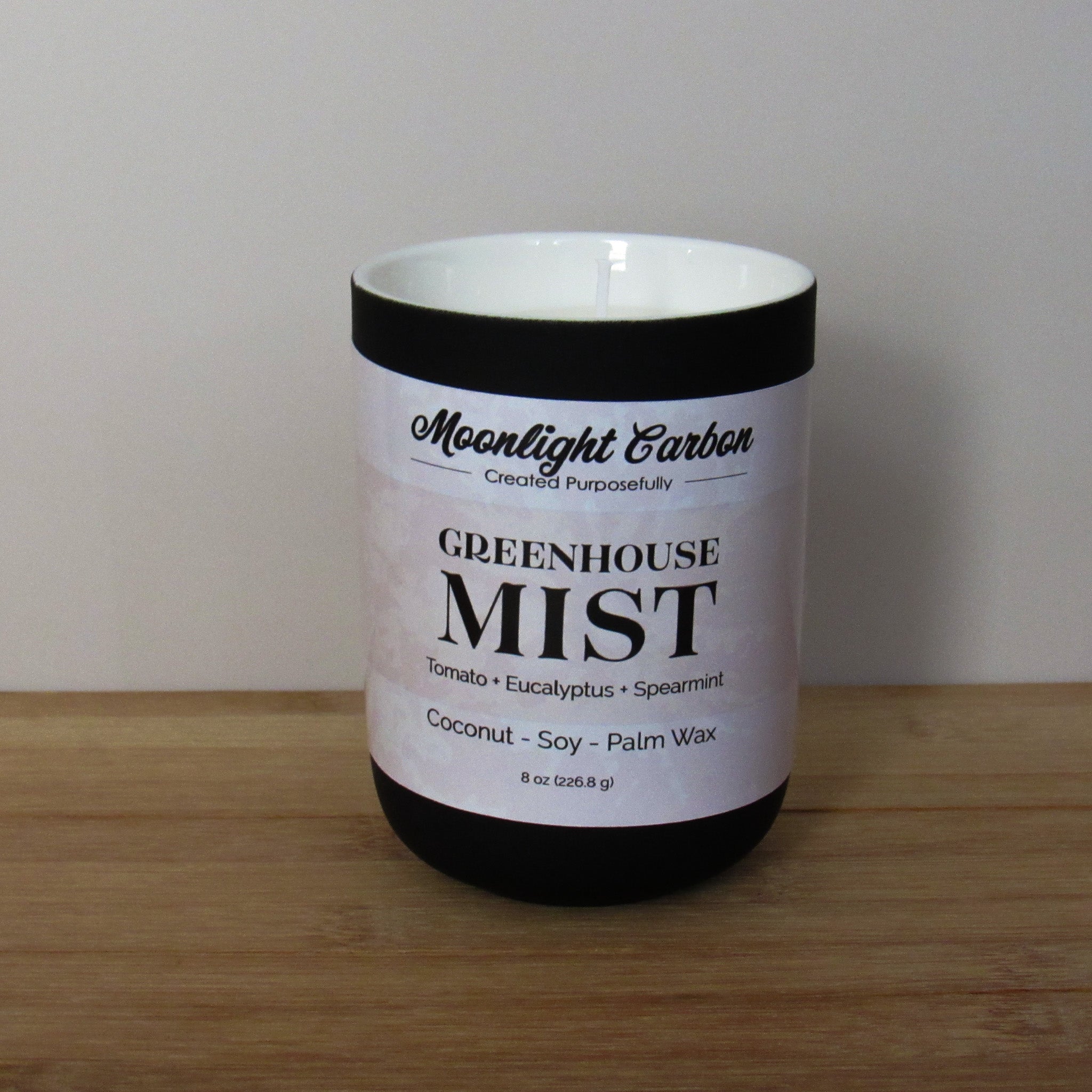 Candle with a labeled 'Greenhouse Mist' on a wooden surface and plain background. Fragrance notes include tomato, eucalptus, and spearmint.