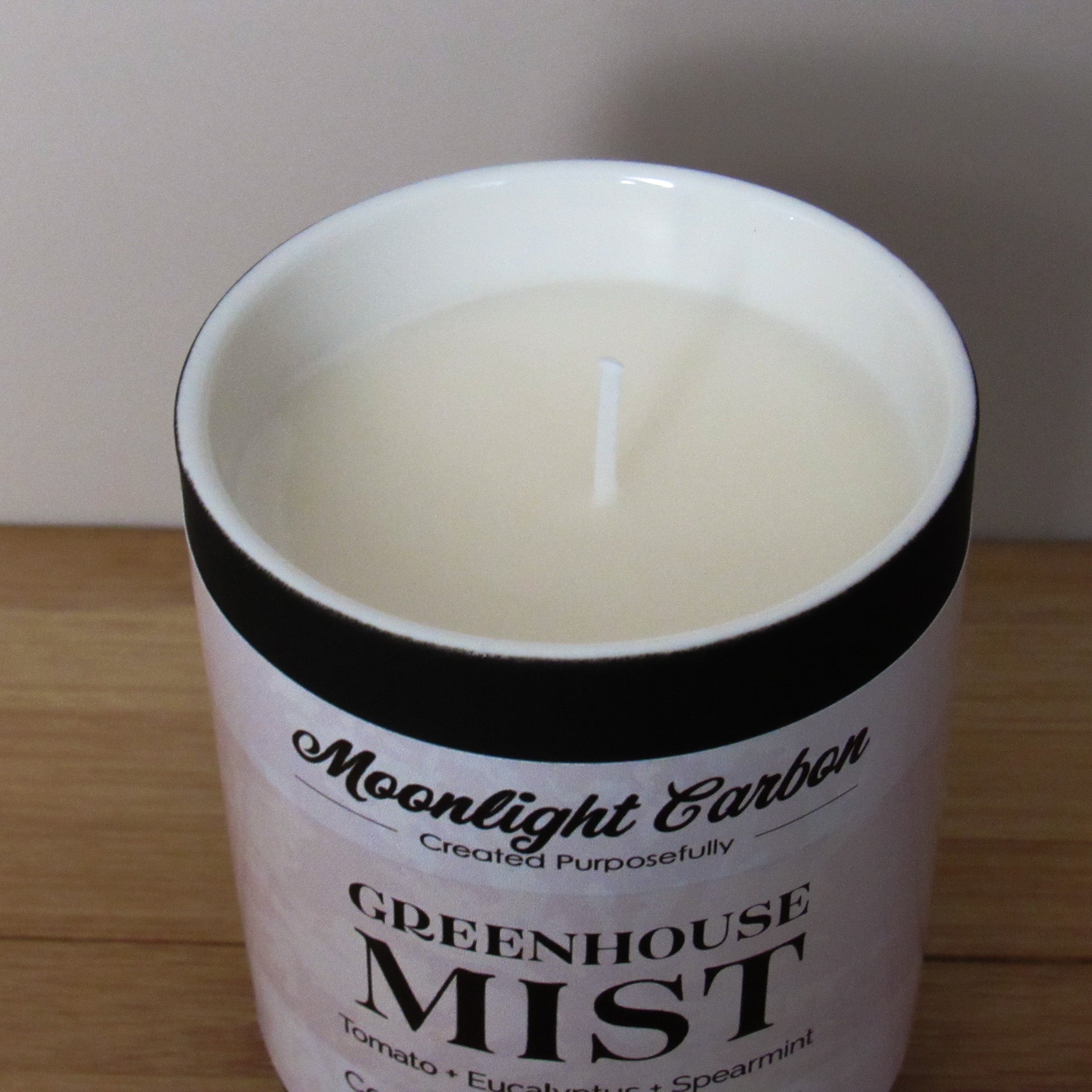 Candle in a container labeled 'Greenhouse Mist' by Moonlight Carbon on a wooden surface. Zoomed into view into top os candle showing coconut, soy, palm wax and cotton wick.