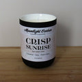 Candle with a labeled 'Crisp Sunrise' on a wooden surface. In a black ceramic jar. Fragrance notes include fresh laundry.