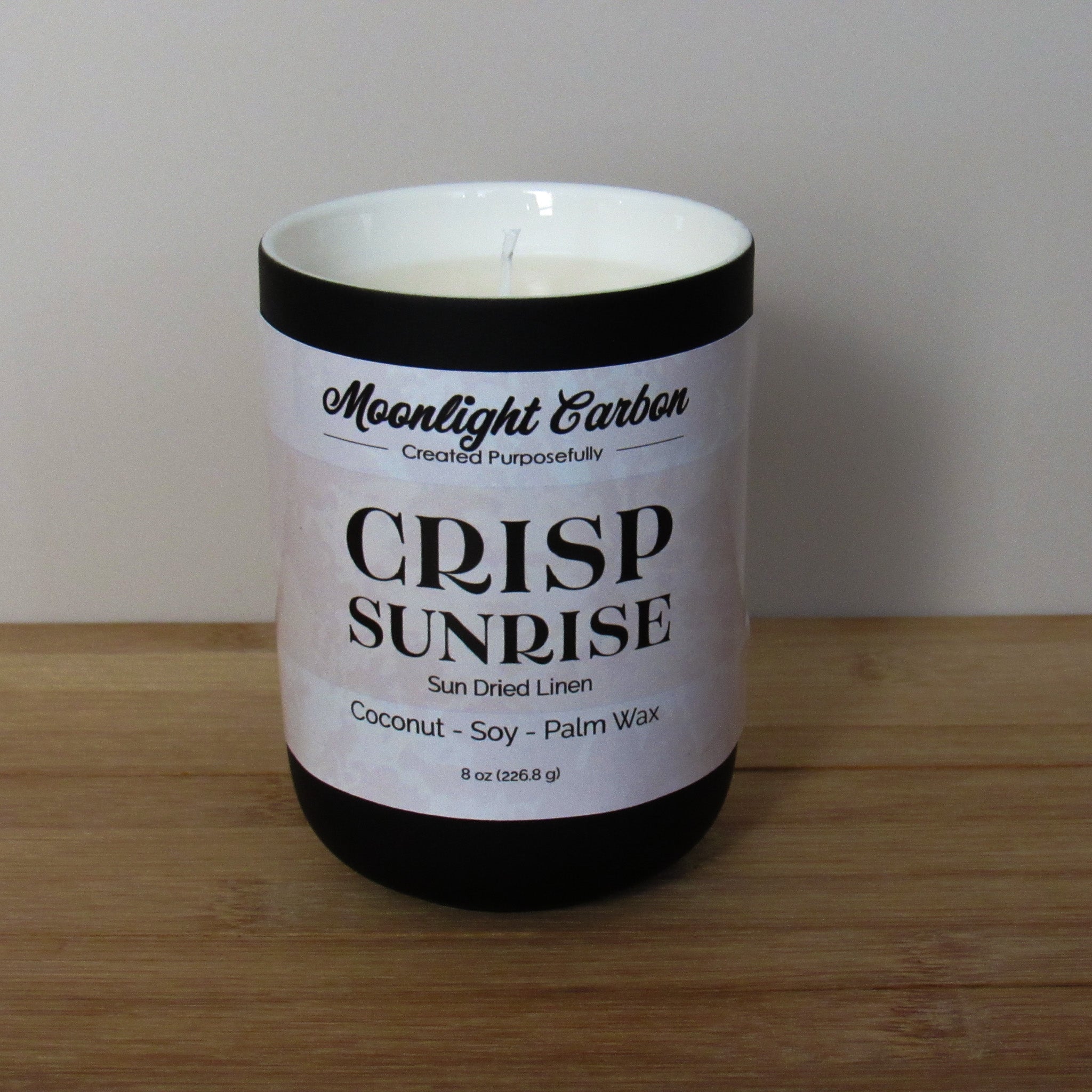 Candle with a labeled 'Crisp Sunrise' on a wooden surface. In a black ceramic jar. Fragrance notes include fresh laundry.