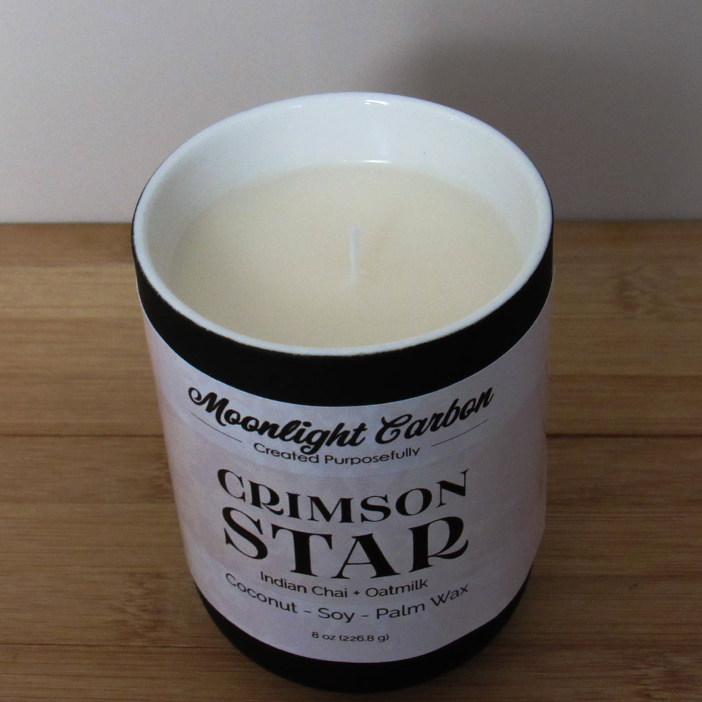 Candle in a black ceramic container with a beige label on a wooden surface. Zoomed into the top of the candle showing coconut, soy palm wax blend and cotton wick.