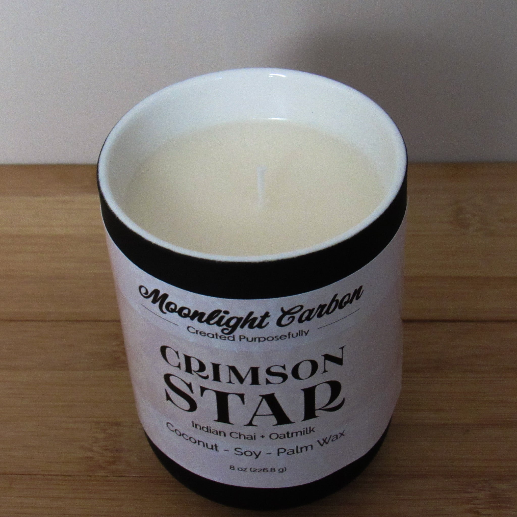 Candle in a black ceramic container with a beige label on a wooden surface. Zoomed into the top of the candle showing coconut, soy palm wax blend and cotton wick.